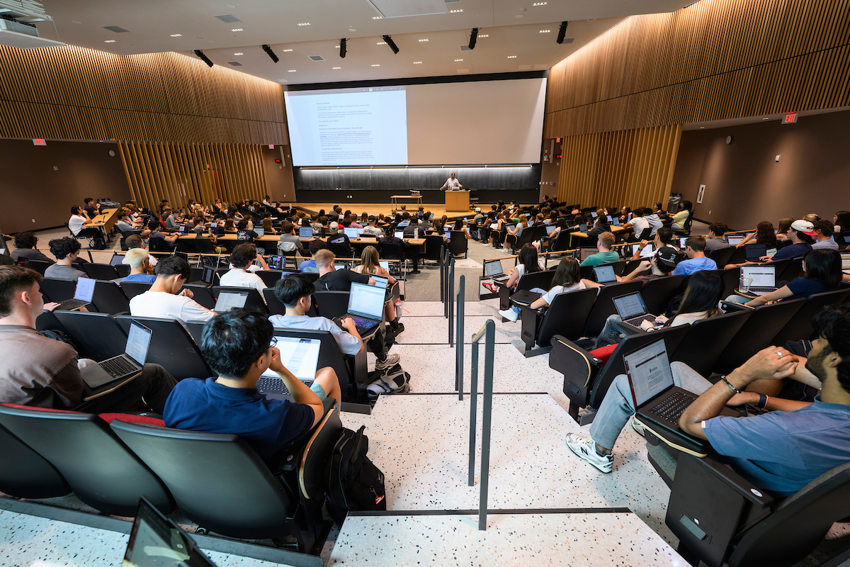 Students with laptops take notes during a large lecture course with an instructor presenting slides 